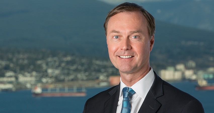 Smiling man in suit and tie stands in front of a cityscape with mountains in the background.